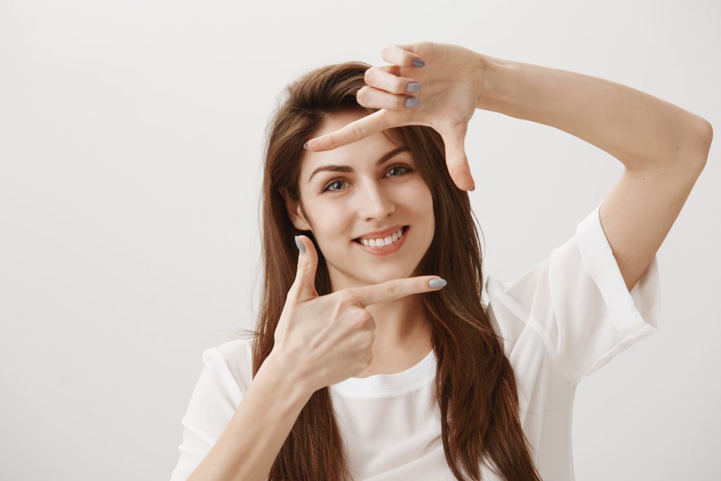 Creative girl imaging how cute her new dress will look on model. Portrait of friendly charming designer finger framing and looking through at camera with broad smile, standing over gray wall.